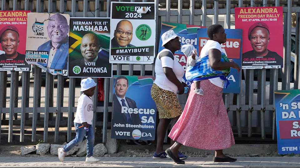 Mulheres com filhos passam por cartazes eleitorais em Tembisa, leste de Joanesburgo, África do Sul, 28 de maio de 2024, antes das eleições de quarta-feira, 29 de maio. Foto: Reprodução People´s World.