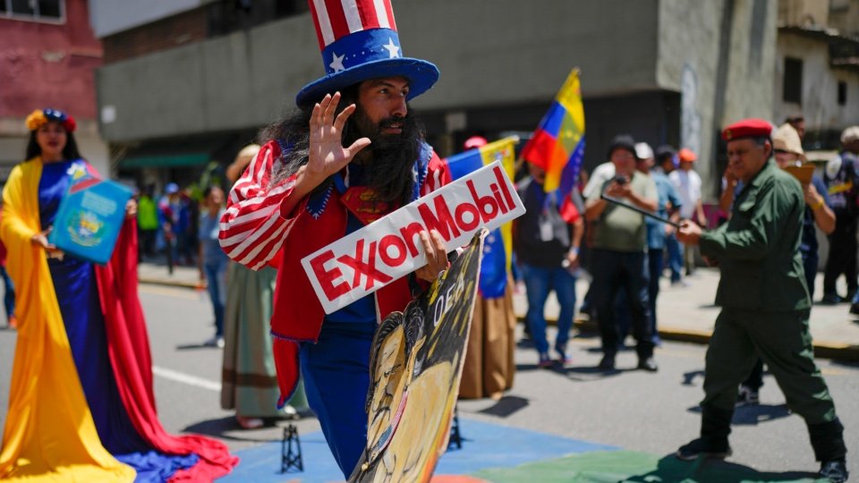 Um venezuelano vestido de Tio Sam segura uma placa da petrolífera ExxonMobil durante uma marcha em Caracas, Venezuela. Para atrair grandes empresas petrolíferas a investir na exploração predatória dos recursos naturais da Venezuela, o presidente Donald Trump está oferecendo dinheiro dos contribuintes americanos para subsidiar os custos da extração. | Ariana Cubillos / AP Um venezuelano vestido de Tio Sam segura uma placa da petrolífera ExxonMobil durante uma marcha em Caracas, Venezuela. Para atrair grandes empresas petrolíferas a investir na exploração predatória dos recursos naturais da Venezuela, o presidente Donald Trump está oferecendo dinheiro dos contribuintes americanos para subsidiar os custos da extração. | Ariana Cubillos / AP